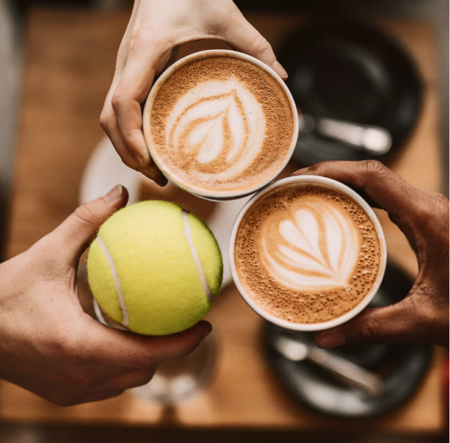 Two hands holding lattes over a table beside a tennis ball